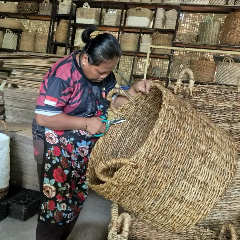 An Indonesian artisan meticulously finishing a large handcrafted woven basket, surrounded by various natural fiber products in the PT. Mitra Eco Bhavana workshop, showcasing sustainable production