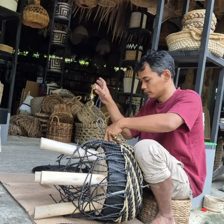 An Indonesian artisan hand-weaving a natural fiber product, possibly a woven lamp or basket, in a workshop filled with various handcrafted rattan and natural goods from PT. Mitra Eco Bhavana